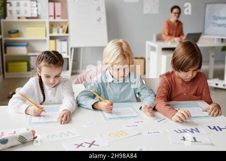 Group of school children sitting at the table and making notes on papers during lesson at school with teacher in the background Stock Photo