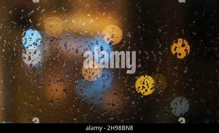 Close-up of the blurred light spots through the window and raindrops falling down on the glass at the evening. Rainy weather Stock Photo