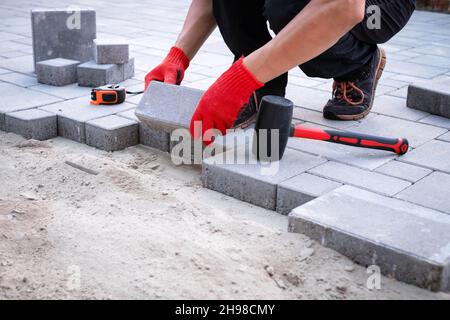 The master in yellow gloves lays paving stones in layers. Garden brick pathway paving by professional paver worker. Laying gray concrete paving slabs in house courtyard on sand foundation base. Stock Photo