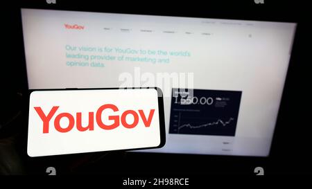 Person holding smartphone with logo of British market research company YouGov Plc on screen in front of website. Focus on phone display. Stock Photo