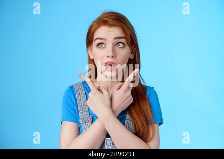 Uncertain cute redhead female making decision, cross arms body, pointing sideways upper left right corners, look aside hesitant, thinking, open mouth Stock Photo