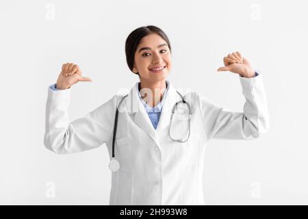 Glad smiling young indian female doctor in coat points fingers at herself, isolated on white background, studio Stock Photo