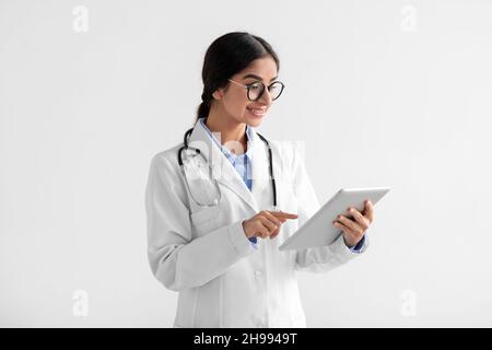 Happy young indian lady doctor in white coat and glasses working on tablet Stock Photo