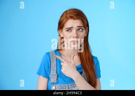 Stupified redhead hesitant girl show aversion, pointing looking left reluctant, full disbelief, stare with dismay uncertain about product, disliking Stock Photo