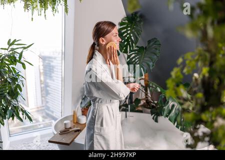 Side view of young beautiful woman exfoliating facial skin with mask remover cellulose face sponge, removing makeup or doing cleansing routine Stock Photo