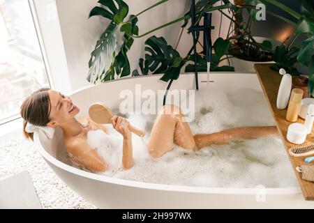 Cheerful young woman holding brush and singing in it while relaxing in bathtub Stock Photo