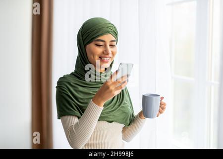 Happy young Arab woman in hijab standing near window with coffee and smartphone, browsing internet, chatting online Stock Photo