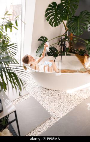 Cheerful young woman having fun while pouring shower gel on loofah sponge, taking bath in modern bathroom decorated with tropical plants Stock Photo