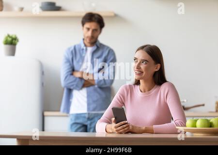 Offended sad millennial male looks at smiling female with smartphone at kitchen Stock Photo