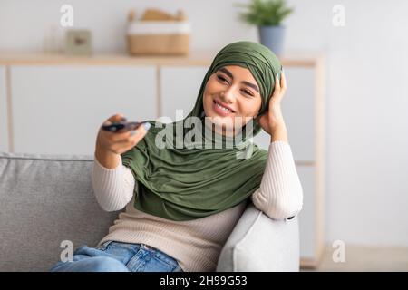 Young Arab woman in Muslim hijab holding remote control, watching TV on sofa at home Stock Photo