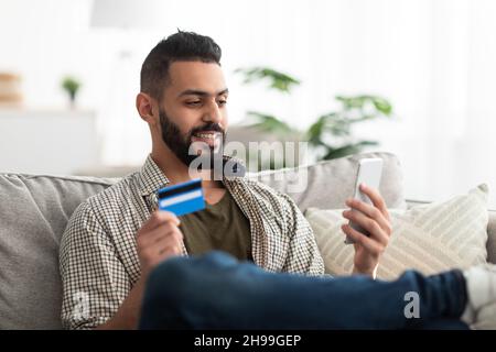 Attractive young Arab man using mobile phone and credit card for online shopping or remote banking at home Stock Photo