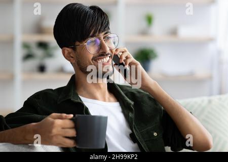 Happy young indian man sitting on sofa, talking on phone Stock Photo
