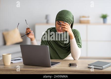 Arab female in hijab feeling tired, rubbing irritated eyes, using laptop, feeling exhausted from online work at home Stock Photo