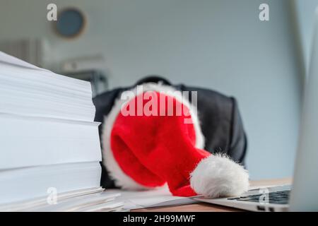 Tired and exhausted businessman with Santa Claus hat fell asleep over his office desk during Christmas season holiday, selective focus Stock Photo