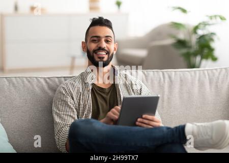 Handsome young Arab man using digital tablet while sitting on couch at home Stock Photo