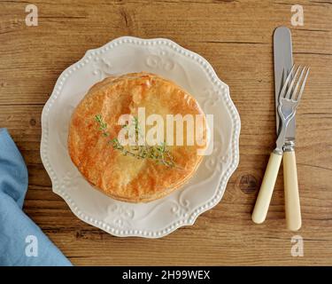 Fresh baked succulent chicken pot pie with vegetables and sprig of thyme in flat lay composition.  Vintage cutlery and blue linen napkin on warm wood Stock Photo