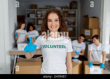 Happy young woman in blue gloves showing thumb up and smiling at camera, working with colleagues in charity center Stock Photo