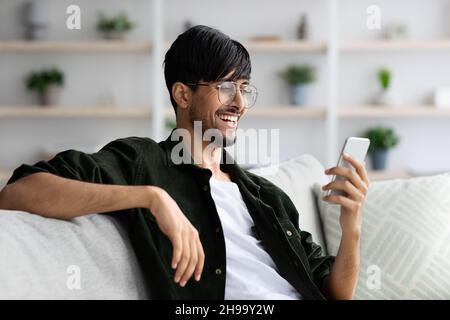 Happy indian guy sitting on couch, using smartphone Stock Photo