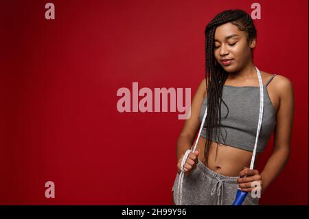 Attractive African woman with stylish dreadlocks and fit body posing against red background with skipping rope. Cardio workout and fitness training co Stock Photo
