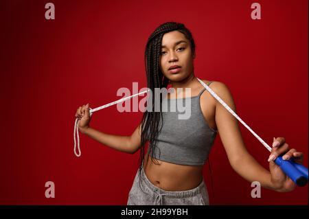 African woman with stylish dreadlocks and fit body posing against red background with jumping rope. Cardio workout, weight loss and fitness training c Stock Photo