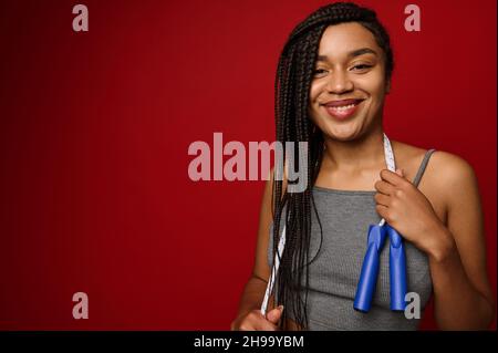 Cheerful African woman with dreadlocks posing against a red background with a skipping rope around her neck, smiling with a toothy smile, looking at t Stock Photo