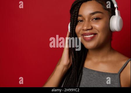 Headshot of an attractive stunning delighted cheerful African young woman with stylish dreadlocks, wearing wireless headphones, smiles toothy smile lo Stock Photo