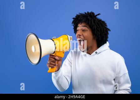 Cool news. Agitated African American teen guy screaming into megaphone over blue studio background Stock Photo