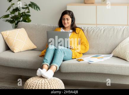 Telecommuting concept. Excited arab woman sitting on sofa and working online on laptop, wearing headset Stock Photo
