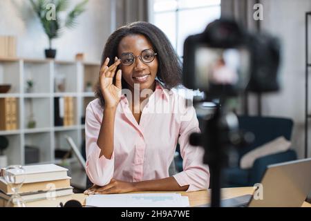Pleasant african american woman in eyeglasses using video camera for online lessons with students. Smiling female teacher sitting at desk with book and notes. Stock Photo