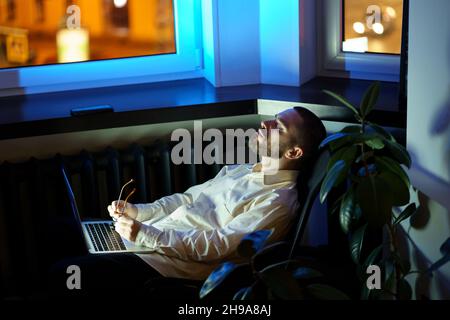 Exhausted young businessman or programmer sit with closed eyes in dark office with laptop overtime Stock Photo