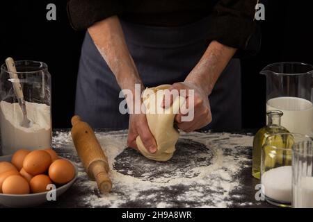 A man baker kneads dough on the table, milk in a jug and flour, chicken eggs in a plate and sugar and butter, male hobby for cooking Stock Photo