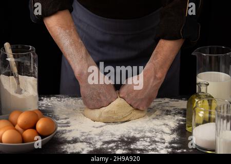 A man baker kneads dough on the table, milk in a jug and flour, chicken eggs in a plate and sugar and butter, male hobby for cooking Stock Photo