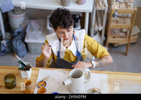 Woman artisian in pottery studio drawing on clay handmade teapot, female ceramist at work Stock Photo
