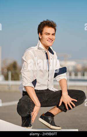 Young man in unfastened white shirt sits on squats on the street Stock Photo