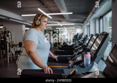 Mid adult overweight woman with headphones exercising on treadmill in gym Stock Photo