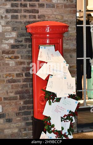 An English red postbox with letters to Father Christmas, Santa ,flowing out of it with a brick wall back drop and holly leaves and berries Stock Photo