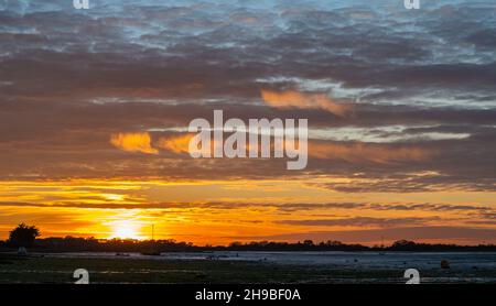 Cloudy orange sky as the sun sets over Bosham Quay in the harbour by Bosham Village at low tide in Winter in West Sussex, England, UK. Stock Photo