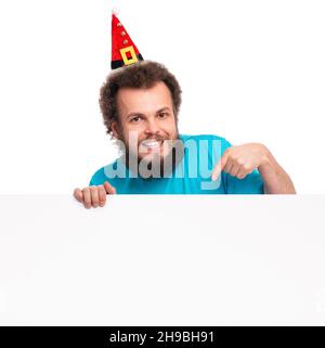 Smiling bearded man wear Santa Claus hat with blank sign, point finger empty space isolated over white background. Stock Photo