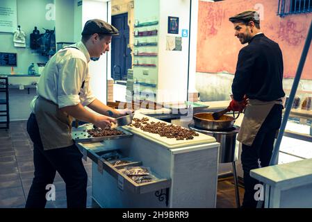 Two young men wearing similar caps making nut sweets in a small shop. Stock Photo