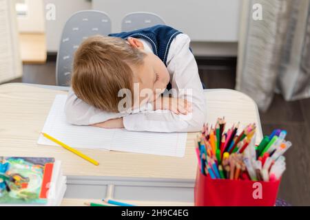 A cute first grader boy in a school uniform at home during a pandemic fell asleep doing homework at a desk with books and pencils. Selective focus. Cl Stock Photo