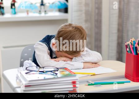 A cute first grader boy in a school uniform at home during a pandemic fell asleep doing homework at a desk with books and pencils. Selective focus. Cl Stock Photo