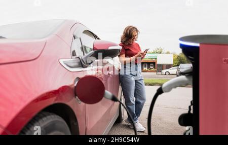Woman with smartphone next to charging red electric car  Stock Photo