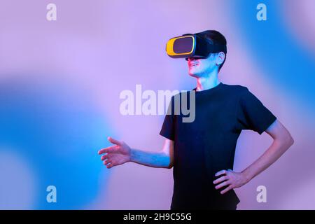A young man in virtual reality glasses gives a hand to say hello. Communication in virtual reality. Neon colors, copy space Stock Photo