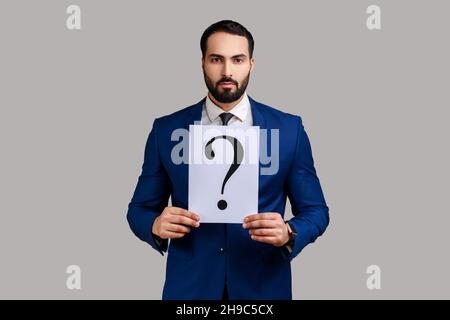 Portrait of serious bearded businessman looking at camera, holding paper with question mark, thinks about tasks, wearing official style suit. Indoor studio shot isolated on gray background. Stock Photo