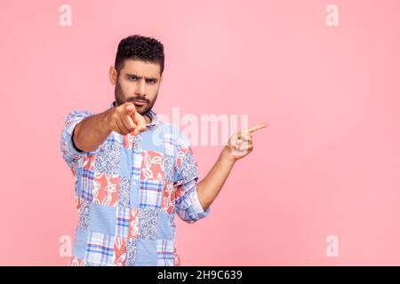 Young adult serious angry man pointing at you with finger and presenting area with promotion, necessarily pay attention, has strict expression. Indoor studio shot isolated on pink background. Stock Photo