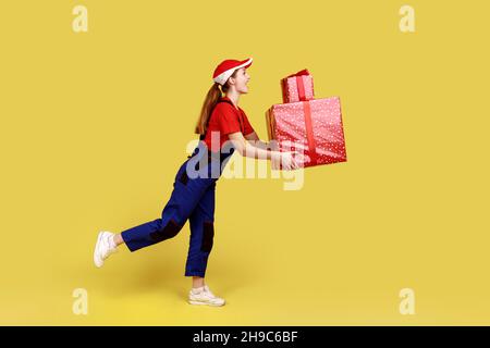 Side view portrait of courier woman being hurry to deliver present boxes for clients, door-to-door delivery, with wearing overalls and red cap. Indoor studio shot isolated on yellow background. Stock Photo