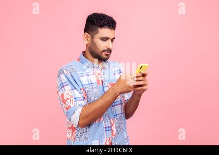 Side view of serious concentrated brunette man with beard holding cell phone in hands, chatting or checking e-mail, gadget addiction. Indoor studio shot isolated on pink background. Stock Photo