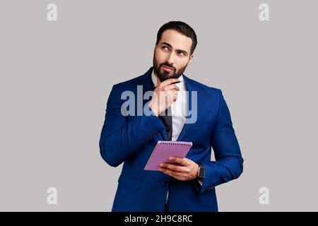 Young adult businessman standing with thoughtful expression, holding paper notebook, pondering business idea, future plans, wearing official style suit. Indoor studio shot isolated on gray background. Stock Photo