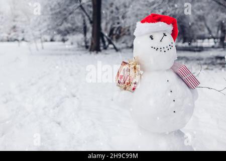 Happy snowman wearing festive Santa hat in snowy winter park at Christmas season outdoors holding presents gift boxes. Winter activities Stock Photo