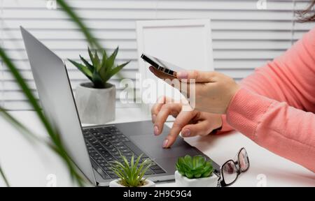a woman sits at a white table holds a smartphone in her hand, with the other hand types on a laptop keyboard. Online shopping, freelance telecommuting Stock Photo